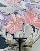 A vibrant mural featuring large, stylized flowers with pink and lavender petals dominates the background. A small, whimsical fairy-like figure is nestled among the petals, adding a fantastical element. In front of the mural, there are two elegant gold chairs with black seats facing a round black table.