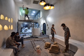 People are working on the interior renovation of a room that appears to be under construction. A worker on a scaffold is painting the ceiling, while others are seated or discussing the progress. The room has smooth, gray walls with arched recesses and a modern design vibe. There are tools and materials scattered around, including a ladder, paint cans, and some rocks or construction debris on the floor. Overhead, stylish pendant lights provide illumination as natural light streams in from windows.