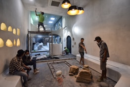 People are working on the interior renovation of a room that appears to be under construction. A worker on a scaffold is painting the ceiling, while others are seated or discussing the progress. The room has smooth, gray walls with arched recesses and a modern design vibe. There are tools and materials scattered around, including a ladder, paint cans, and some rocks or construction debris on the floor. Overhead, stylish pendant lights provide illumination as natural light streams in from windows.