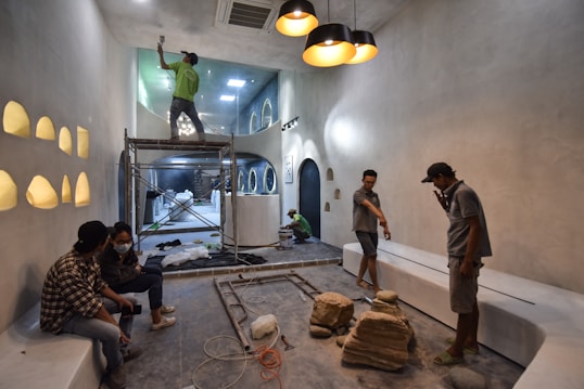 People are working on the interior renovation of a room that appears to be under construction. A worker on a scaffold is painting the ceiling, while others are seated or discussing the progress. The room has smooth, gray walls with arched recesses and a modern design vibe. There are tools and materials scattered around, including a ladder, paint cans, and some rocks or construction debris on the floor. Overhead, stylish pendant lights provide illumination as natural light streams in from windows.