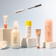 A collection of skincare and cosmetic products is arranged on white blocks against a soft blue background. The products vary in size and packaging, including tubes and bottles labeled with the brand name 'Glossier'. A mascara wand and a small tube are suspended above the arrangement, adding a dynamic element to the composition.