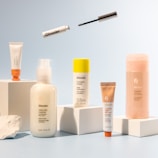A collection of skincare and cosmetic products is arranged on white blocks against a soft blue background. The products vary in size and packaging, including tubes and bottles labeled with the brand name 'Glossier'. A mascara wand and a small tube are suspended above the arrangement, adding a dynamic element to the composition.