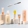 A collection of skincare and cosmetic products is arranged on white blocks against a soft blue background. The products vary in size and packaging, including tubes and bottles labeled with the brand name 'Glossier'. A mascara wand and a small tube are suspended above the arrangement, adding a dynamic element to the composition.