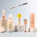 A collection of skincare and cosmetic products is arranged on white blocks against a soft blue background. The products vary in size and packaging, including tubes and bottles labeled with the brand name 'Glossier'. A mascara wand and a small tube are suspended above the arrangement, adding a dynamic element to the composition.
