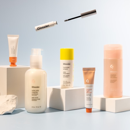 A collection of skincare and cosmetic products is arranged on white blocks against a soft blue background. The products vary in size and packaging, including tubes and bottles labeled with the brand name 'Glossier'. A mascara wand and a small tube are suspended above the arrangement, adding a dynamic element to the composition.