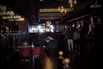 A dimly lit bar or lounge with several people standing and sitting around. The space is furnished with plush chairs and tables, and a bar area with bottles is visible in the background. Warm lighting from chandeliers and pendant lights creates a cozy atmosphere.