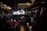 A dimly lit bar or lounge with several people standing and sitting around. The space is furnished with plush chairs and tables, and a bar area with bottles is visible in the background. Warm lighting from chandeliers and pendant lights creates a cozy atmosphere.