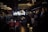 A dimly lit bar or lounge with several people standing and sitting around. The space is furnished with plush chairs and tables, and a bar area with bottles is visible in the background. Warm lighting from chandeliers and pendant lights creates a cozy atmosphere.