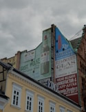 Advertising murals painted on the side of old buildings, featuring products and store names with large typography against a cloudy sky. The building facades include muted colors with architectural details.