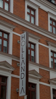 A multi-story brick building with large windows framed by white moldings. The prominently featured vertical sign reads 'Hollandia'. The structure displays a classic architectural style with red brickwork and decorative elements.