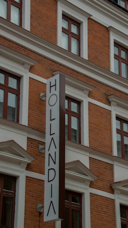 A multi-story brick building with large windows framed by white moldings. The prominently featured vertical sign reads 'Hollandia'. The structure displays a classic architectural style with red brickwork and decorative elements.