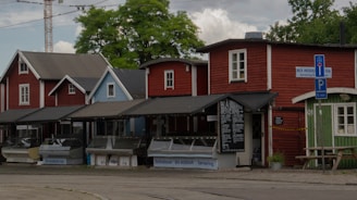 A row of charming wooden buildings with red and blue facades, featuring slanted roofs typical of Nordic architecture. These houses are fronts for small businesses, one of which is a seafood shop with a blue sign. A few outdoor seating areas with protective awnings are present, along with a wooden sign advertising various seafood offerings. Potted plants add a touch of greenery to the scene, which is framed by lush green trees and a cloudy sky.