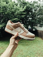 A person is holding a sneaker in an outdoor setting with greenery in the background. The sneaker has a white base with brown suede overlays and a white Nike logo on the side.