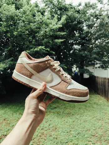 A person is holding a sneaker in an outdoor setting with greenery in the background. The sneaker has a white base with brown suede overlays and a white Nike logo on the side.