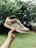 A person is holding a sneaker in an outdoor setting with greenery in the background. The sneaker has a white base with brown suede overlays and a white Nike logo on the side.