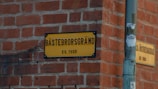 A yellow street sign with black letters is mounted on a red brick wall at a corner. The text reads 'BÄSTEBRORSGRÄND KV. THOR'. Next to the street sign, there is a pole with a sticker that has some graphic and text on it.