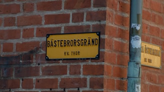 A yellow street sign with black letters is mounted on a red brick wall at a corner. The text reads 'BÄSTEBRORSGRÄND KV. THOR'. Next to the street sign, there is a pole with a sticker that has some graphic and text on it.