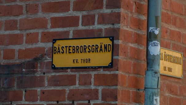 A yellow street sign with black letters is mounted on a red brick wall at a corner. The text reads 'BÄSTEBRORSGRÄND KV. THOR'. Next to the street sign, there is a pole with a sticker that has some graphic and text on it.