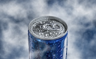 A close-up view of a blue beverage can, covered in water droplets. The background is softly blurred in shades of gray and white, providing a cool, refreshing feel.