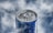A close-up view of a blue beverage can, covered in water droplets. The background is softly blurred in shades of gray and white, providing a cool, refreshing feel.