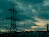 A large, metal electricity pylon stands against a moody sky filled with dark clouds. Power lines extend from the pylon, stretching across the horizon. Below, there are silhouettes of buildings, adding an urban element to the scene.