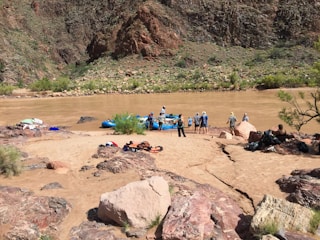 A group of people prepare for rafting on a muddy river, surrounded by rocky terrain and sparse vegetation. Brightly colored equipment like life jackets and rafts are scattered on the sandy shore.