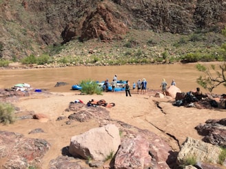 A group of people prepare for rafting on a muddy river, surrounded by rocky terrain and sparse vegetation. Brightly colored equipment like life jackets and rafts are scattered on the sandy shore.