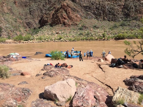 A group of people prepare for rafting on a muddy river, surrounded by rocky terrain and sparse vegetation. Brightly colored equipment like life jackets and rafts are scattered on the sandy shore.