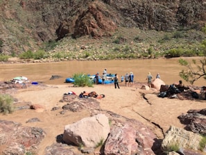 A group of people prepare for rafting on a muddy river, surrounded by rocky terrain and sparse vegetation. Brightly colored equipment like life jackets and rafts are scattered on the sandy shore.