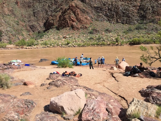 A group of people prepare for rafting on a muddy river, surrounded by rocky terrain and sparse vegetation. Brightly colored equipment like life jackets and rafts are scattered on the sandy shore.