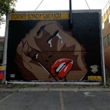 A mural depicting a close-up of a face with striking red lips, tears streaming from closed eyes, and hands held up to the face. The artwork includes the text 'SORRY IS NOT ENOUGH...' in bold yellow letters at the top, with 'BLACK LIVES MATTER' and 'DENIAL' written on the right side.