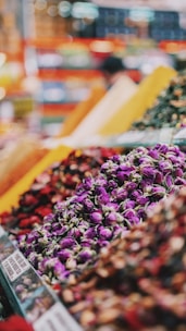 A vibrant market scene features a variety of colorful spices and dried flowers organized in large piles. The foreground showcases purple rosebuds, while warm tones of reds, yellows, and greens create a lively and aromatic atmosphere. Blurred backgrounds suggest bustling activity.