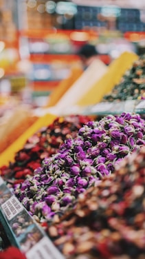 A vibrant market scene features a variety of colorful spices and dried flowers organized in large piles. The foreground showcases purple rosebuds, while warm tones of reds, yellows, and greens create a lively and aromatic atmosphere. Blurred backgrounds suggest bustling activity.