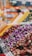 A vibrant market scene features a variety of colorful spices and dried flowers organized in large piles. The foreground showcases purple rosebuds, while warm tones of reds, yellows, and greens create a lively and aromatic atmosphere. Blurred backgrounds suggest bustling activity.