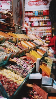 An organized display of various colorful spices and herbs in a vibrant marketplace. The spices are neatly arranged in rows, each with a distinctive color and texture, creating a visually appealing pattern. A large banner with 'Hazer Baba' is visible in the background, emphasizing a focus on quality.