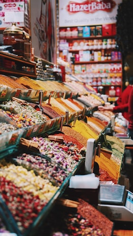 An organized display of various colorful spices and herbs in a vibrant marketplace. The spices are neatly arranged in rows, each with a distinctive color and texture, creating a visually appealing pattern. A large banner with 'Hazer Baba' is visible in the background, emphasizing a focus on quality.