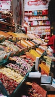 An organized display of various colorful spices and herbs in a vibrant marketplace. The spices are neatly arranged in rows, each with a distinctive color and texture, creating a visually appealing pattern. A large banner with 'Hazer Baba' is visible in the background, emphasizing a focus on quality.