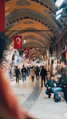 A bustling indoor market with arched ceilings adorned with intricate patterns and Turkish flags hanging prominently. People are walking through the corridor, some browsing the various shops displaying colorful textiles, lamps, and other goods.