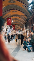 A bustling indoor market with arched ceilings adorned with intricate patterns and Turkish flags hanging prominently. People are walking through the corridor, some browsing the various shops displaying colorful textiles, lamps, and other goods.