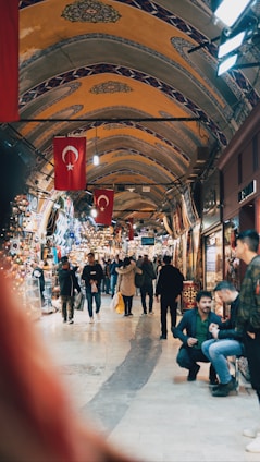 A bustling indoor market with arched ceilings adorned with intricate patterns and Turkish flags hanging prominently. People are walking through the corridor, some browsing the various shops displaying colorful textiles, lamps, and other goods.