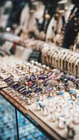 A display of an assortment of rings and bracelets is showcased within a glass case. The rings feature various gemstones in diverse colors including purple, red, and blue, while the bracelets are lined up in the background. The setting conveys an elegant atmosphere with the sparkling and intricate designs of the jewelry.