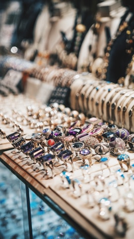 A display of an assortment of rings and bracelets is showcased within a glass case. The rings feature various gemstones in diverse colors including purple, red, and blue, while the bracelets are lined up in the background. The setting conveys an elegant atmosphere with the sparkling and intricate designs of the jewelry.