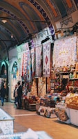 A well-lit market stall showcasing a variety of colorful rugs and textiles displayed prominently. Intricate patterns and vibrant hues adorn the walls and tables, creating an inviting atmosphere. Arched ceilings with elaborate designs contribute to the aesthetic appeal. People are seen browsing and possibly engaged in conversation.