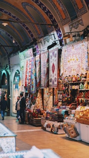 A well-lit market stall showcasing a variety of colorful rugs and textiles displayed prominently. Intricate patterns and vibrant hues adorn the walls and tables, creating an inviting atmosphere. Arched ceilings with elaborate designs contribute to the aesthetic appeal. People are seen browsing and possibly engaged in conversation.