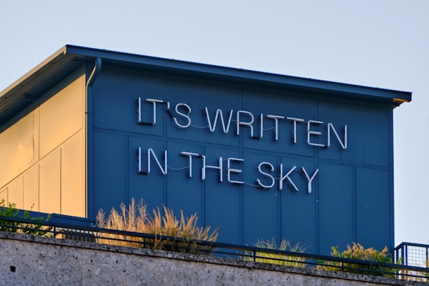 A blue-painted building rooftop with the phrase 'IT'S WRITTEN IN THE SKY' displayed in white neon lights. The structure features some vegetation along the edge.