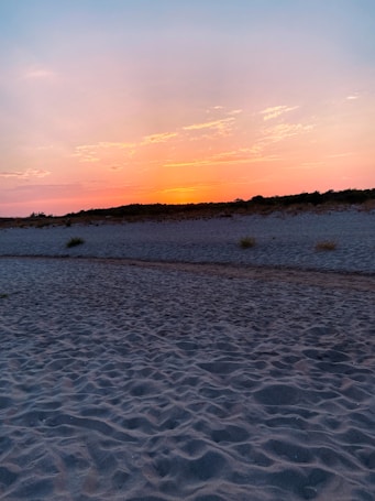The image captures a serene beach landscape at sunset. The sky displays a gradient of colors ranging from soft pinks and oranges to a gentle blue. The sun is partially visible near the horizon, casting a warm glow across the scene. The sandy beach is in the foreground, leading to low dunes and sparse vegetation in the background.