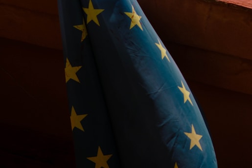 A dark scene featuring part of a flag with yellow stars on a blue field, indicative of the European Union flag. The flag is hanging, with visible wrinkles and shadows creating a moody atmosphere.