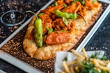A flatbread topped with spicy chicken, green peppers, and garnished with fresh scallions on a wooden platter. There is a side dish of seasoned fries and a wine glass present in the background, all placed on a dark textured surface.