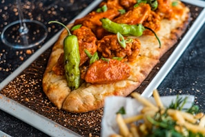 A flatbread topped with spicy chicken, green peppers, and garnished with fresh scallions on a wooden platter. There is a side dish of seasoned fries and a wine glass present in the background, all placed on a dark textured surface.