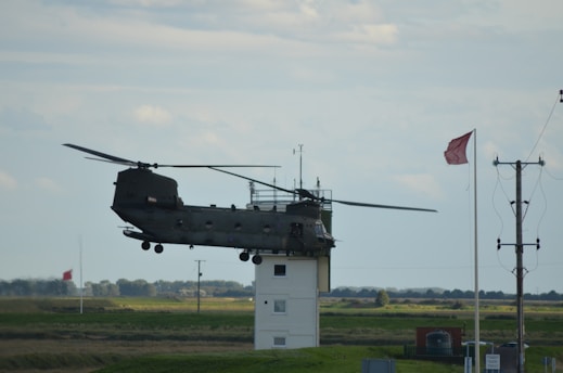 A helicopter is positioned very close to a small building, appearing to be hovering slightly above or in contact with the roof. The background shows a grassy landscape and electric poles. Two red flags are visible, indicating possible wind direction.